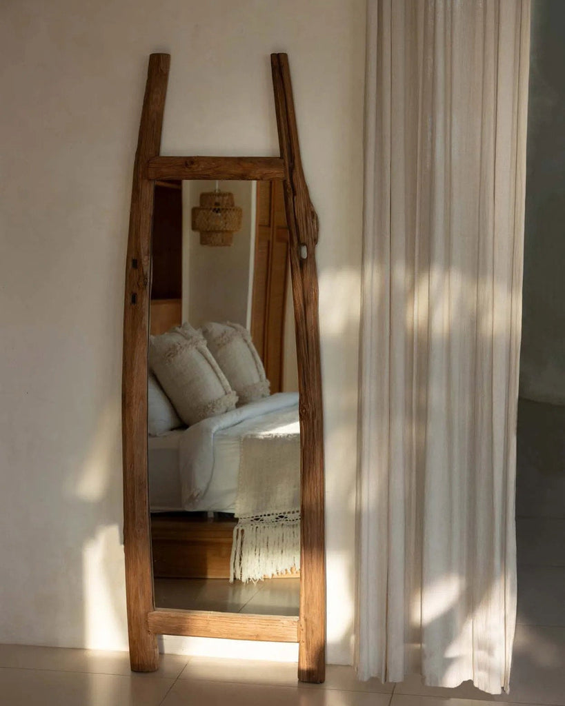 Wooden floor mirror leaning against a wall with a bed and curtains in the background.