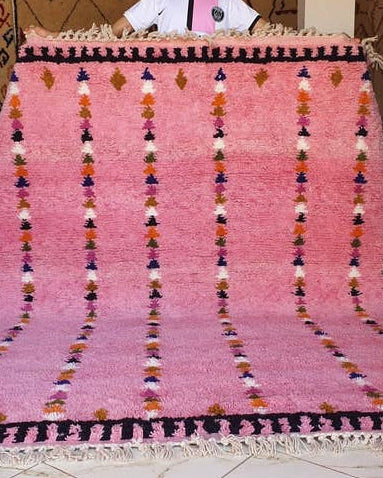 Person holding a large pink rug with decorative patterns in a room.