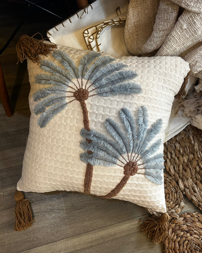 A square-shaped cushion cover with a palm tree design, featuring blue and beige colors, made of cotton, displayed on a chair.