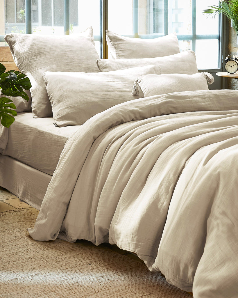 Neatly made bed with beige bedding in a bright room with large windows.